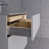 Stilfull badrumsinspiration med beige tvättställsskåp med innerskiva, överskåp, öppen hylla, ovalt spegel med belysning, matchande pendellampor, hylla under spegeln och vägghängd toalett. Väggar med ljusa, avlånga kakelplattor