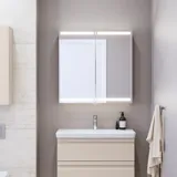 Liten badrumsinspiration med tvättställsskåp och överskåp över toalett i beige specialfärg, elegant spegelskåp med integrerat ljus, de ljusa färgerna och vita gardinen skapar en romantisk och feminin atmosfär.