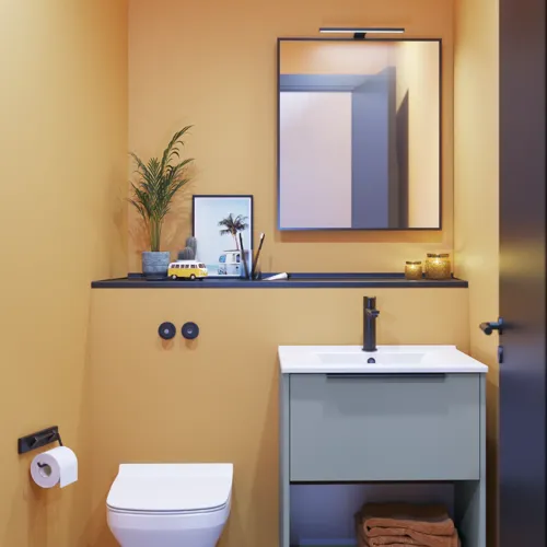Liten badrumsinspiration med sneda och varma gula väggar, elegant tvättställsskåp i ljusblått, tunn och stilren handfat samt minimalistisk fyrkantig spegel med svart kant och praktisk lampa.